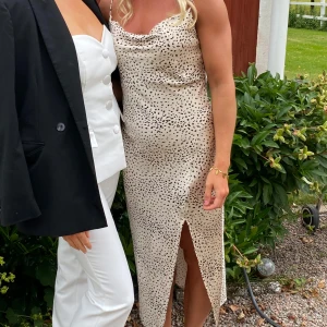 Beige prickig långklänning med slits - Säljer en beige långklänning med svarta prickar och smala axelband. Klänningen har en snygg slits framtill och ett draperat dekolletage. Materialet är glansigt och känns lyxigt, perfekt för dig som vill sticka ut med en trendig look. Fårn Gina 