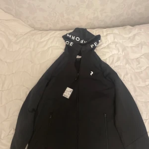 Svart zip hood jacka Peak Performance - Svart zip hood jacka från Peak Performance med vit logga på bröstet och tryckt text på huvan. Jackan har dragkedja framtill och två fickor med dragkedja. Perfekt för dig som gillar sportig och clean stil. Finns alla storlekar