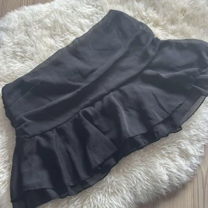 Svart volangkjol  - Supersnygg svart kortkjol med volangkant. Kjolen har en omlott-look och sitter tajt i midjan, perfekt för att skapa en trendig och feminin siluett. Passar dig som gillar att sticka ut med stilrena detaljer och flowy material.