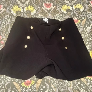 Svarta shorts med guldknappar River Island - Stilrena svarta shorts från River Island med sex snygga guldknappar framtill. Klassisk och lite dressad look med hög midja och kortare ben. Perfekta för dig som vill ha en chic vibe och gillar detaljer som sticker ut.