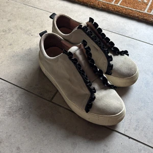 Beige sneakers med svarta rosetter - Snygga beige sneakers i mocka med unika svarta rosett-detaljer längs snörningen. Skorna har platt vit sula och rund tå, vilket ger en cool och trendig look. Perfekta för dig som vill sticka ut med en twist på klassiska sneakers.