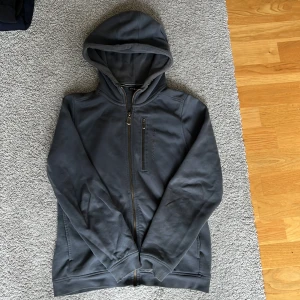 Blå hoodie från Sail Racing - Blå hoodie från Sail Racing med hel dragkedja, bröstficka med dragkedja och broderad logga. Tröjan har huva, långa ärmar och är tillverkad i mjukt bomullsmaterial. Tröjan är använd ganska mycket och har små tecken på det. Hör av dig vid frågor👏