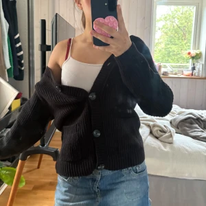 Svart stickad kofta med knappar - Mysig svart stickad kofta med grov ribbstickning och stora knappar framtill. Koftan har två fickor och långärmade ärmar, perfekt att slänga över en topp eller t-shirt. Enkel och stilren design som funkar till jeans eller kjol.