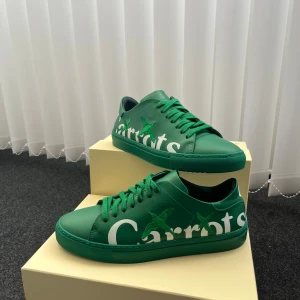 Gröna Axel Arigato Carrots sneakers - Collab mellan Carrots by Anwar Carrots och Axel Arigato. Limited edition. Vit text och broderade gröna fåglar på sidan. Skorna har snörning, platt sula och är tillverkade i skinn. Perfekt för dig som vill sticka ut med en unik och färgstark look. 