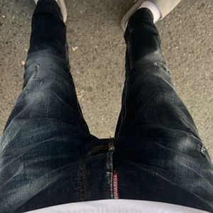 Mörkblå Dsquared2 jeans med slitningar - Säljer ett par mörkblå jeans från Dsquared2 med coola slitningar och tvättade detaljer. Jeansen har en rak passform och klassisk femficksdesign. Röd Dsquared2-logga syns vid gylfen. Perfekta för dig som gillar streetstyle.