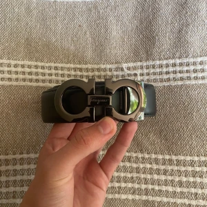 Svart Ferragamo skärp med metallspänne - Snyggt svart skärp från Ferragamo i äkta skinn med ikoniskt, blankt metallspänne i silverfärg. Spännet har Ferragamo-loggan ingraverad och en unik, rundad design som sticker ut. Perfekt accessoar för att lyfta din outfit med en lyxig touch.