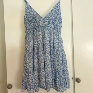 Ljusblå blommig klänning - Supersöt ljusblå kort klänning med vita blommor och volangdetaljer. Klänningen har v-ringad hals och tunna axelband. Bra kvalitet och i storlek S. 