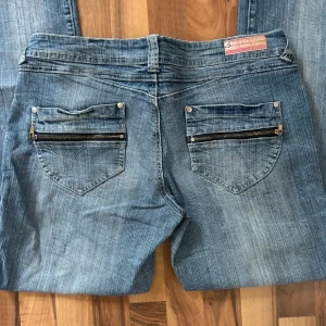 Blå jeans med dragkedjor på bakfickorna - Snygga blå jeans med coola dragkedjor på bakfickorna och klassisk femficksdesign. Jeansen har låg midja och raka ben, tillverkade i mjukt denimtyg. Perfekta för en avslappnad streetstil.