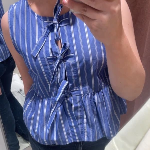 Blåvit randig ärmlös blus med knyt - Superfin blå och vit randig blus med tre knyt framtill och volang nedtill. Ärmlös modell med rund halsringning och luftig passform, perfekt för varma dagar. Gjord i mjuk bomull som känns skön mot huden. Snygg till både jeans och kjol!