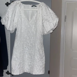 Vit kortklänning med puffärmar - Supersöt vit kortklänning från Gina med puffiga kortärmar och v-ringad hals. Klänningen sitter tajt/är figursydd. Materialet känns mjukt och har en lätt struktur. Använd en gång!