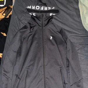 Svart hoodie från Perform - Svart hoodie med huva från Perform. Hoodien har hel dragkedja framtill, två fickor med dragkedja och vit logga på bröstet. På insidan av huvan står det 'PERFORM' i vitt. Perfekt för dig som gillar sportig och clean stil.