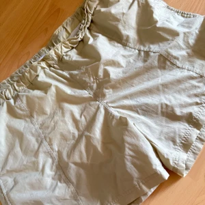 Beige bad/sport shorts med meshfoder och snörning - Snygga beige shorts med elastisk midja och mörkblå snörning. Insidan har vitt meshfoder för extra komfort och ventilation. Liten broderad logga på benet. Perfekta för varma dagar och sportiga aktiviteter.