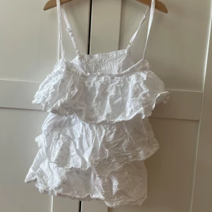 Vit volangig linnetopp med broderi - Supersöt vit linnetopp med flera lager volanger och broderade detaljer. Justerbara smala axelband och smockad rygg för skön passform. Perfekt för sommarens varma dagar och ger en romantisk vibe med sina broderade blommor och luftiga känsla.