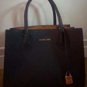 Mörk handväska från Michael Kors - Snygg svart handväska från Michael Kors i skinn med både handtag och axelrem. Väskan har guldfärgade detaljer. Säljer för den inte kommer till användning. Pris går att diskutera.