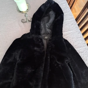 Svart fusk pälsjacka med huva från ONLY - Mysig svart pälsjacka från ONLY i storlek S men passar för någon i storlek 34-36. Jackan har en fluffig känsla, huva och är helsvart. Perfekt för kalla dagar när du vill vara både varm och snygg. 