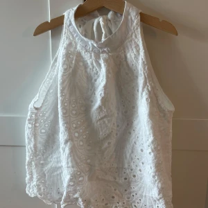 Vit broderad linnetopp med knyt - Superfin vit linnetopp med broderade detaljer och hålmönster. Toppen har hög hals och knytning i nacken för en snygg look. Materialet är lätt och luftigt, perfekt för varma dagar. Passformen är loose och toppen är ärmlös.