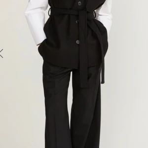 Svart ullväst med bälte från STOCKH LM - Stilren svart väst från STOCKH LM i ull med knappar framtill, två bröstfickor och tillhörande bälte i midjan. Västen har klassisk krage och en rak, något loose passform. Perfekt att slänga över en skjorta för en clean look.