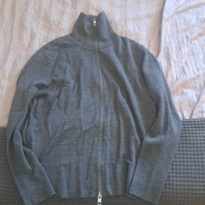 Grå kofta med dragkedja från H&M - Säljer en stilren grå kofta från H&M i storlek S. Tröjan har hög krage, hel dragkedja framtill och är långärmad. Materialet är mjukt och känns skönt mot huden, perfekt för lager-på-lager under hösten och vintern.  Hör av dig i pm vid intresse.