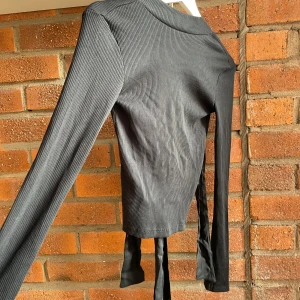 Svart ribbad topp med öppen rygg och knyt - Säljer en svart ribbad långärmad topp i storlek XS med öppen rygg och långa band för knytning. Materialet är mjukt och stretchigt, perfekt för dig som gillar detaljer som sticker ut. Snygg att matcha med jeans eller kjol för en cool look.