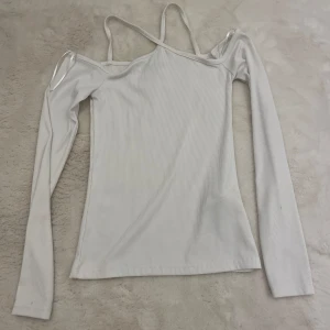 Vit topp - Trendig vit långärmad topp med ribbad struktur och offshoulder-design. Snygga tunna axelband ger en cool twist och gör toppen extra unik. Perfekt att styla med jeans eller kjol för en fräsch look.