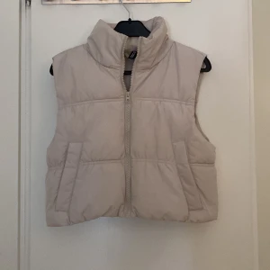 Beige dunväst från H&M Divided XS - Snygg beige dunväst från H&M Divided i storlek XS. Västen har hög krage, två fickor framtill och stängs med dragkedja. Perfekt att slänga över en hoodie för en chill look. Enkel och stilren design som passar till det mesta.