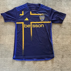 Boca Juniors tröja - Snygg Boca Juniors fotbollströja i mörkblått med gula detaljer och tryck. Adidas-logga på bröstet, klubbmärke och sponsortryck fram och bak. Tillverkad i lätt och ventilerande material, perfekt för träning eller match. Borta tröjan från förra året.HELT NY!