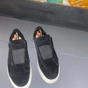 Svarta sneakers från Axel Arigato - Snygga svarta sneakers från Axel Arigato i mocka med elastisk panel över vristen och vit platt sula. Minimalistisk design med diskret logga i guld på sidan. Perfekta för dig som gillar stilrena och trendiga skor. Storleken sitter lite mindre än en vanlig 41, så passar bättre om du har 39-40,5 i storlek 