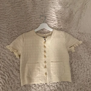 Beige blus med volangärm och fickor - Supersöt beige blus i strukturerat tyg med korta ärmar och volangdetaljer. Framtill finns dekorativa guldknappar och två fickor. Blusen har en rak passform och känns både chic och trendig, perfekt till jeans eller kjol. Bara använd en gång.