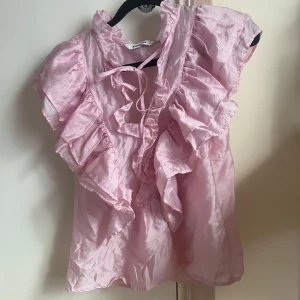 Rosa volangblus med knyt från KappAhl - Supersöt ljusrosa blus från KappAhl med stora volanger längs axlar och bröst. Blusen har korta ärmar och knytning vid halsen för extra detalj. Tillverkad i ett glansigt, tunt material som känns lyxigt och luftigt.