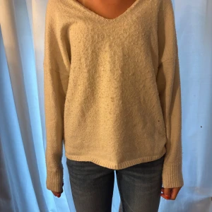 Beige stickad v-ringad tröja - Mysig beige stickad tröja med v-ringning och långa ärmar. Tröjan har en avslappnad passform och ribbade muddar vid armslutänden.
