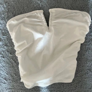 Vit bandeau tubtopp med v-ringning - Säljer en stilren vit bandeau tubtopp med snygg v-ringning framtill. Toppen är ärmlös och har lätt rynkade sidor för en schysst passform. Perfekt för sommardagar eller när du vill ha en enkel men trendig look.