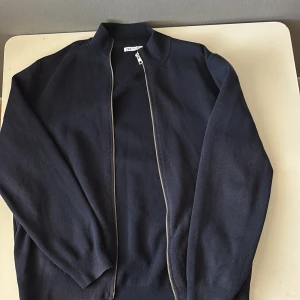 Mörkblå stickad bomberjacka från Zara - Snygg mörkblå stickad bomberjacka från Zara med hel dragkedja framtill och ribbade muddar. Jackan har en clean look med ståkrage och lång ärm, perfekt för dig som gillar stilren och enkel design.