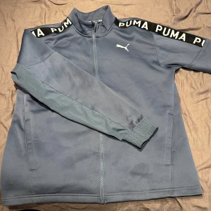 Blå träningsjacka från Puma  - Snygg blå träningsjacka från Puma med dragkedja och hög krage. Svarta axelband med vit PUMA-logga ger en sportig vibe. Jackan har långa ärmar, ribbade muddar och två fickor framtill. Perfekt för träning eller chill dagar. Obs en liten fett fläck där fram men det går säkert att tvätta bort enkelt! Tröjan va en present t min pappa men tyvärr va den för stor så säljer den nu! Vi tror fläcken va där när vi köpte den😅För den är bara använd en gång!  Pris går att diskutera😇💕 hör gärna av er vid frågor
