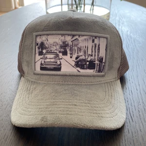 Ljus grå keps från Emilio Pendenza - Snygg ljus grå keps från Emilio Pendenza med mesh på sidorna och baksidan för extra ventilation. Framtill finns en unik patch med svartvit bild av en stadsscen med bil och cyklist. Kepsen har böjd skärm och cool streetstil, perfekt för dig som gillar detaljer.