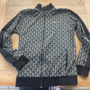 Svart och grå crewneck från Lacoste - Säljer en snygg svart och grå crewneck från Lacoste med hel dragkedja och ribbade muddar. Jackan har ett geometriskt mönster över hela ytan och den klassiska Lacoste-loggan på bröstet. Perfekt för dig som vill sticka ut med stilren streetwear.