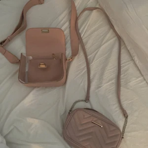 Två rosa axelväskor med kedjedetaljer - Säljer två  axelväskor i ljusrosa. Ena väskan har quiltad design med kedja på axelremmen och dragkedja upptill. Den andra är mer boxig med guldiga detaljer och justerbar rem, samt en tillhörande liten necessär. Perfekt för dig som gillar stilrena accessoarer. 200kr för väskan i andra bilden. 100kr för väskan i första bilden. Pris kan diskuteras. 