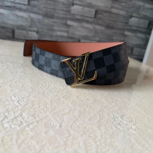 Louis Vuitton rutig skinnbälte - Snyggt Louis Vuitton bälte i svart och grått rutigt mönster med ikonisk LV-spänne i guld. Bältet är tillverkat i äkta skinn med brun insida och har exklusiva detaljer som Louis Vuitton-gravyr på spännet. Perfekt för dig som vill ha en lyxig accessoar med statement.