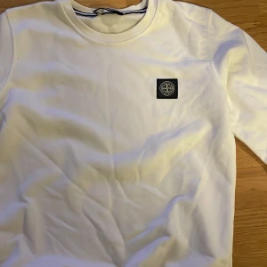 Vit långärmad tröja Stone Island. Den har ingen stone Island märke på armen - Snygg vit långärmad tröja från Stone Island med klassisk rund hals och ikonisk logopatch på bröstet. Tröjan har en clean look och är gjord i mjuk bomull, perfekt för dig som gillar stilrena plagg med streetkänsla. 