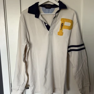 Beige pikétröja med blå detaljer - Snygg beige långärmad pikétröja från Polo Ralph Lauren med mörkblå krage och två blå ränder på ena ärmen. Stor gul P-applikation och siffrorna 67 på bröstet. Perfekt för en sportig och avslappnad stil.
