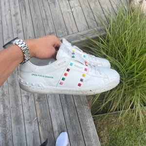 Valentino Rockstud sneakers med färgglada nitar - Snygga vita Valentino Rockstud sneakers med coola färgglada nitar längs sidorna. Skorna har texten 'OPEN FOR A CHANGE' på sidan och är tillverkade i skinn med platt sula och snörning. Perfekta för dig som vill sticka ut med en unik och trendig look. 