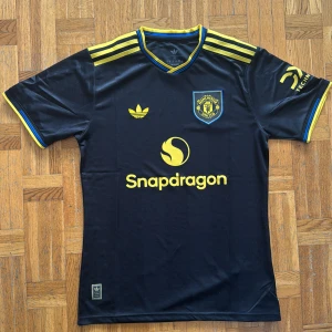 Manchester United fotbollströja - Helt ny! Snygg svart Manchester United fotbollströja från Adidas med gula detaljer och loggor. Tröjan har gula ränder på axlarna, klubbmärke och sponsortryck i gult samt blå inslag vid krage och ärmslut. Tillverkad i lätt och ventilerande polyester.Storleken är XL men passar även L!