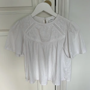 Vit broderad blus från Zara - Supersöt vit blus från Zara med broderade detaljer och spets framtill. Blusen har korta puffärmar, rund halsringning och knapp i nacken. Perfekt för dig som gillar romantisk och luftig stil. Tröjan har även en liten defekt på baksidan som visas på sista bilden❤️