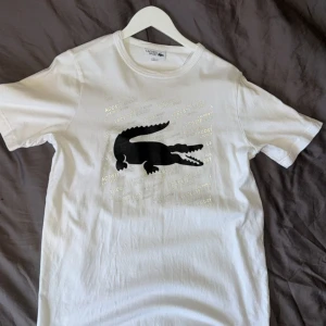Lacoste t-shirt med tryck - Vit t-shirt från Lacoste Sport i bomull med klassisk rund hals och korta ärmar. Stor svart krokodiltryck på bröstet och diskret Lacoste Sport-text i bakgrunden. Liten broderad krokodil på nederkanten. Perfekt för dig som gillar sportig och clean stil.