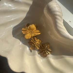 Guldiga blommiga örhängen och ring - Säljer ett set med guldiga smycken i form av blommor. Setet innehåller ett par örhängen och en ring, båda med detaljerade blomblad och glansig finish. Perfekt för dig som vill sticka ut med en cool och unik stil. Blommönstret ger en romantisk och trendig vibe.