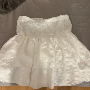 Vit volangkjol med smock - Supersöt vit kortkjol med bred smock i midjan och volanger. Kjolen har en lätt glansig struktur och är perfekt för sommarens alla tillfällen. Den är luftig och har en feminin känsla med sin volangkant.