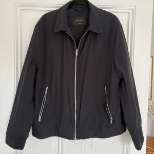 Marinblå vindjacka från Massimo Dutti L - Stilren svart vindjacka från Massimo Dutti med klassisk krage och silverfärgade dragkedjor i Storlek L. Knappt använd. Jackan har två snedställda fickor med dragkedja och enkel design som passar till många outfits. Materialet är lätt och smidigt, perfekt för blåsiga dagar.