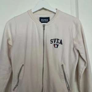 Beige kofta med dragkedja från Svea - Snygg beige kofta från Svea i bomull med broderad logga på bröstet och två fickor med dragkedja. Jackan har rund halsringning och stängs med dragkedja framtill. 