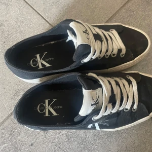 Svarta sneakers från Calvin Klein Jeans - Svarta sneakers från Calvin Klein Jeans med vit logga på sidan och plös. Skorna har vita snören och vit, randig sula. Tillverkade i textil med klassisk låg profil och rund tå! Storlek 38