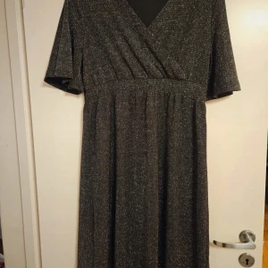 Glittrig svart klänning Mywear Woman - Säljer en svart klänning från Mywear Woman i storlek S. Klänningen har glittriga detaljer, omlott-v-ringning och korta ärmar. Materialet är polyester med metallfiber och elastan, vilket ger en snygg stretch och skimrande look. Perfekt för dig som vill sticka ut.