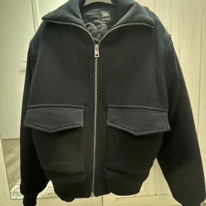 Svart bomberjacka från Zara, M - Snygg svart bomberjacka från Zara med stora fickor framtill och dragkedja hela vägen upp. Jackan har en klassisk krage och ribbade muddar vid ärmslut och nederkant. Perfekt för dig som gillar en stilren och enkel look. Endast använt en gång 